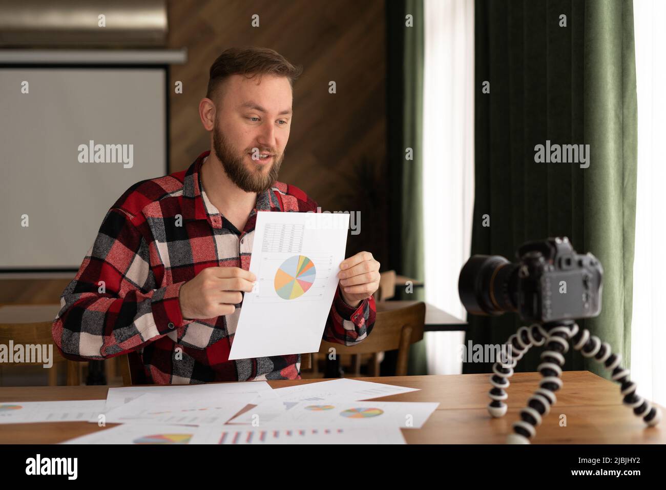 Hombre en camisa mostrar estadísticas gráfico pad hacer vídeo promocional o sesión de fotos en la videocámara a trípode. Vlogger selfie solución de venta o asesor financiero Foto de stock