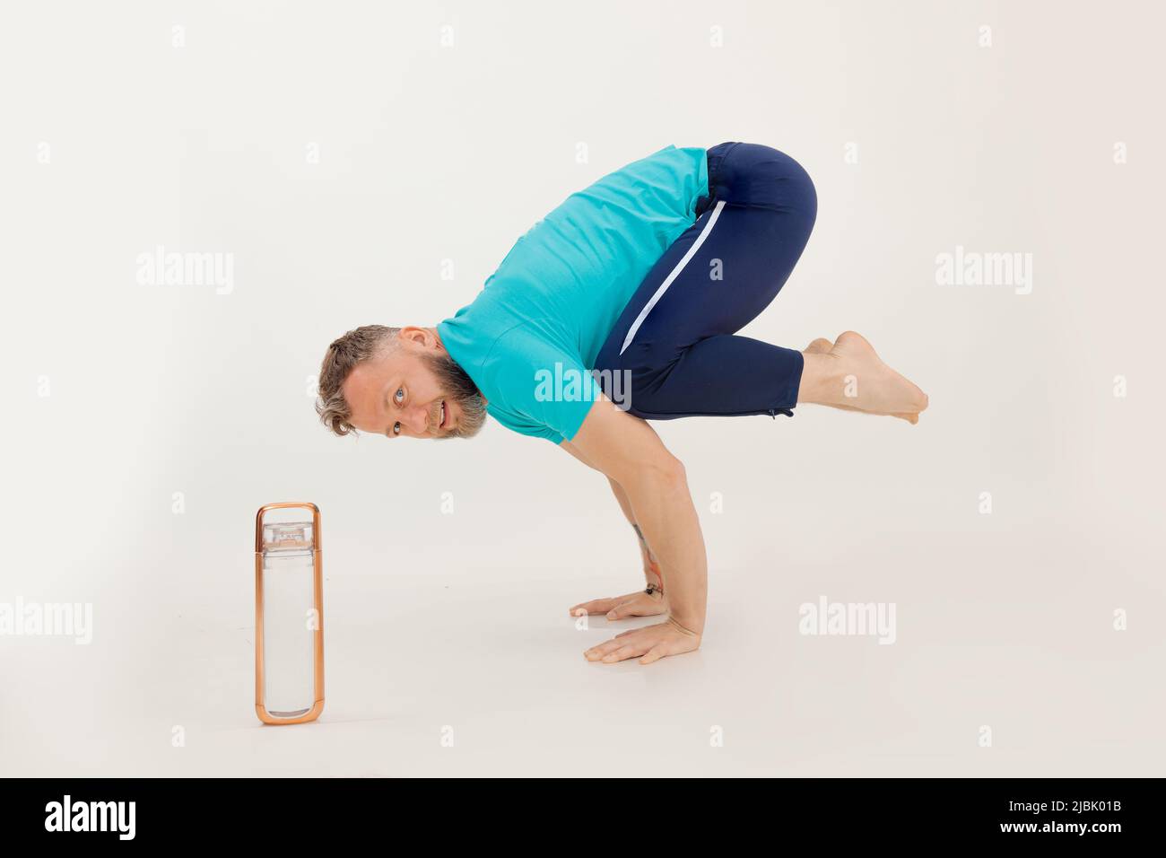 Joven atleta de pie en soporte acrobático con piernas dobladas cerca de una botella de agua, fondo blanco. Mantenerse en forma por el ejercicio en el gimnasio Foto de stock