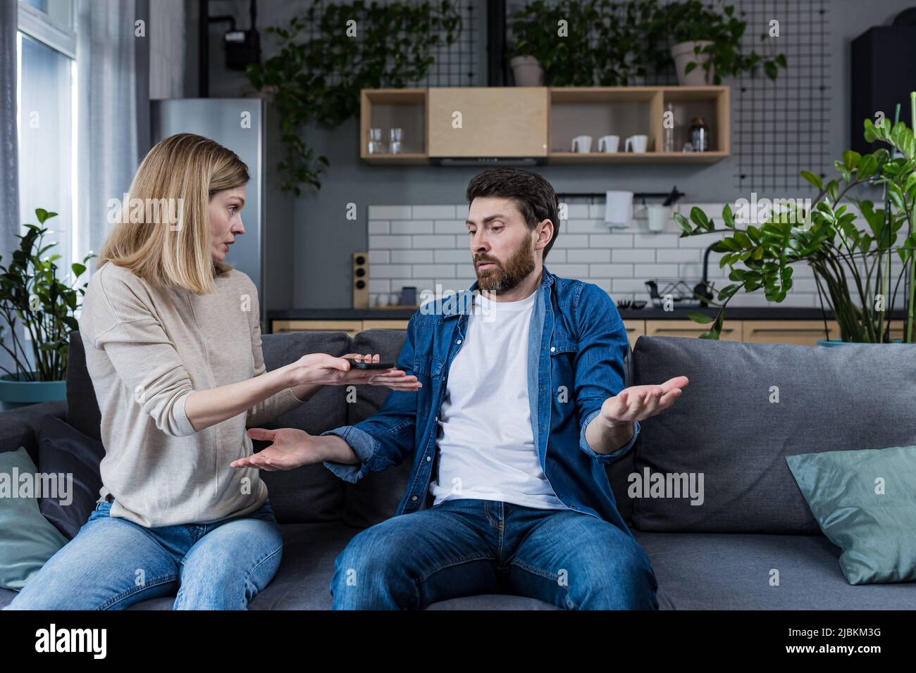 Una mujer disputa con su marido, la reclamación de un hombre perezoso que gana poco dinero, una pareja casada disputa sentado en el sofá Foto de stock