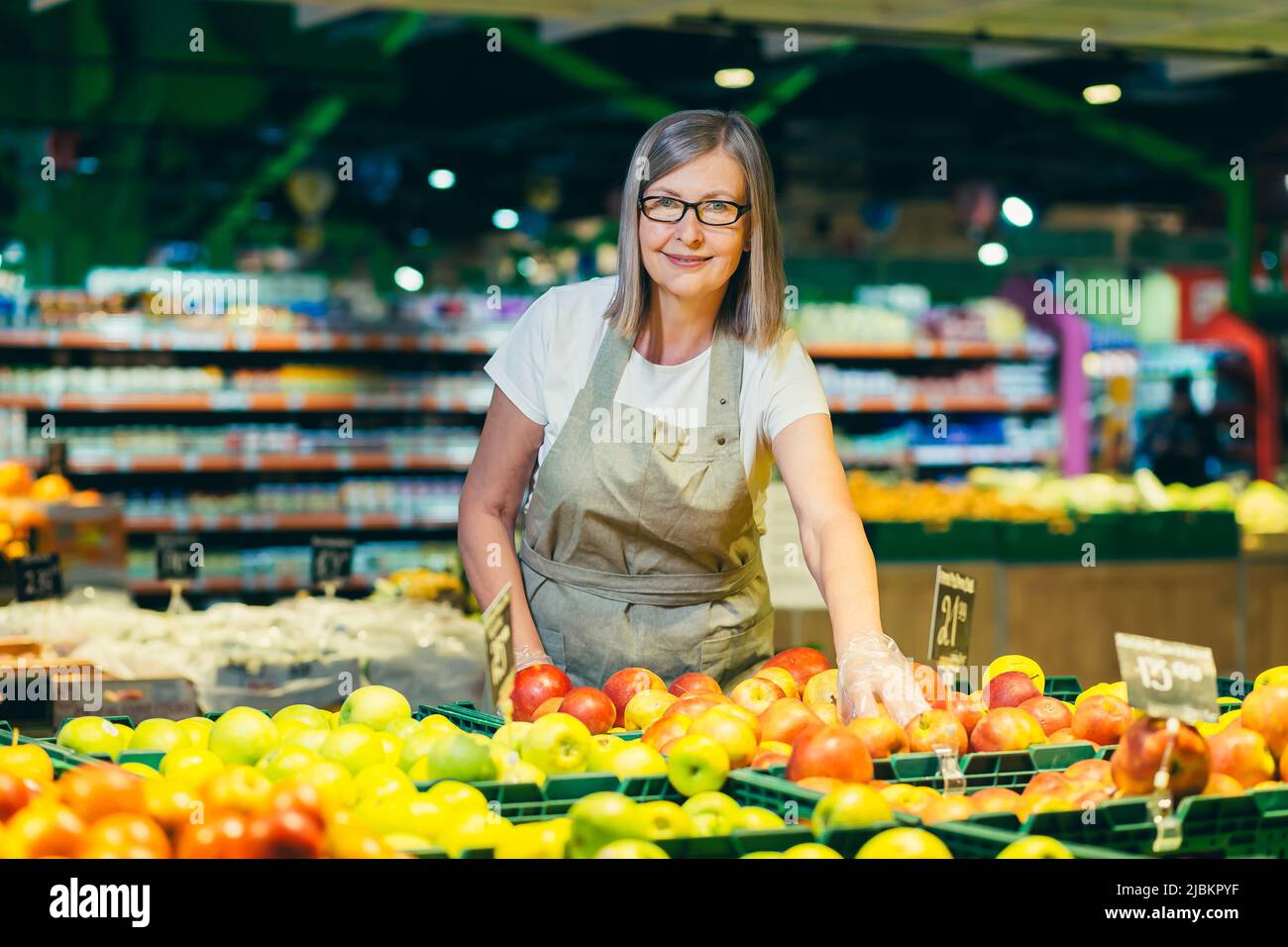 Retrato de una mujer mayor en gafas en la tienda de comestibles empleado,  supermercado. Se coloca en la ropa de trabajo, cerca de frutas y ofochi,  muestra la comida, mira la c
