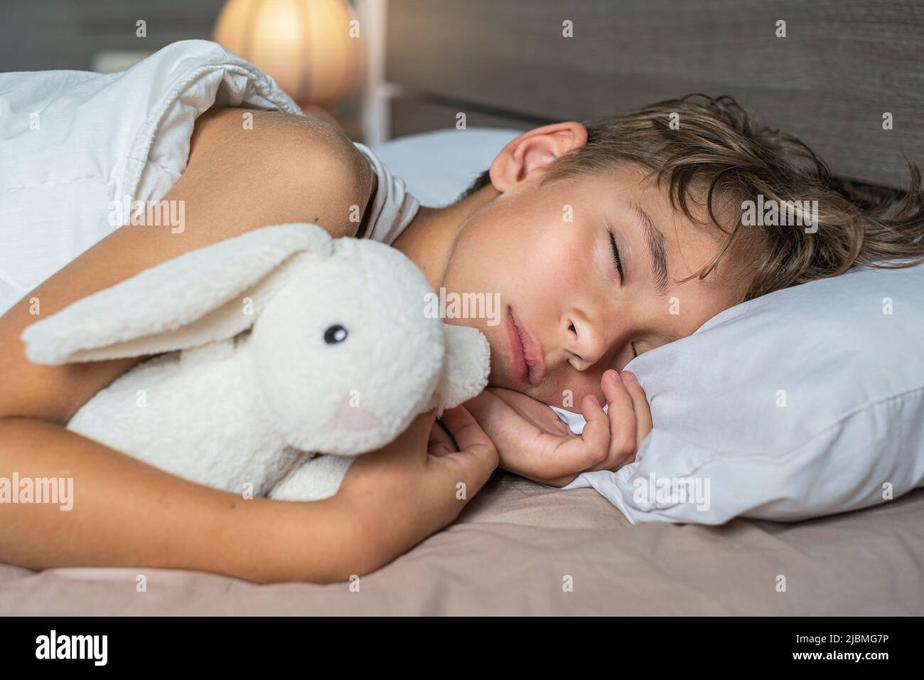 lindo chico durmiendo en la cama con lámpara. Niño de la escuela soñando y sosteniendo juguete de peluche Foto de stock