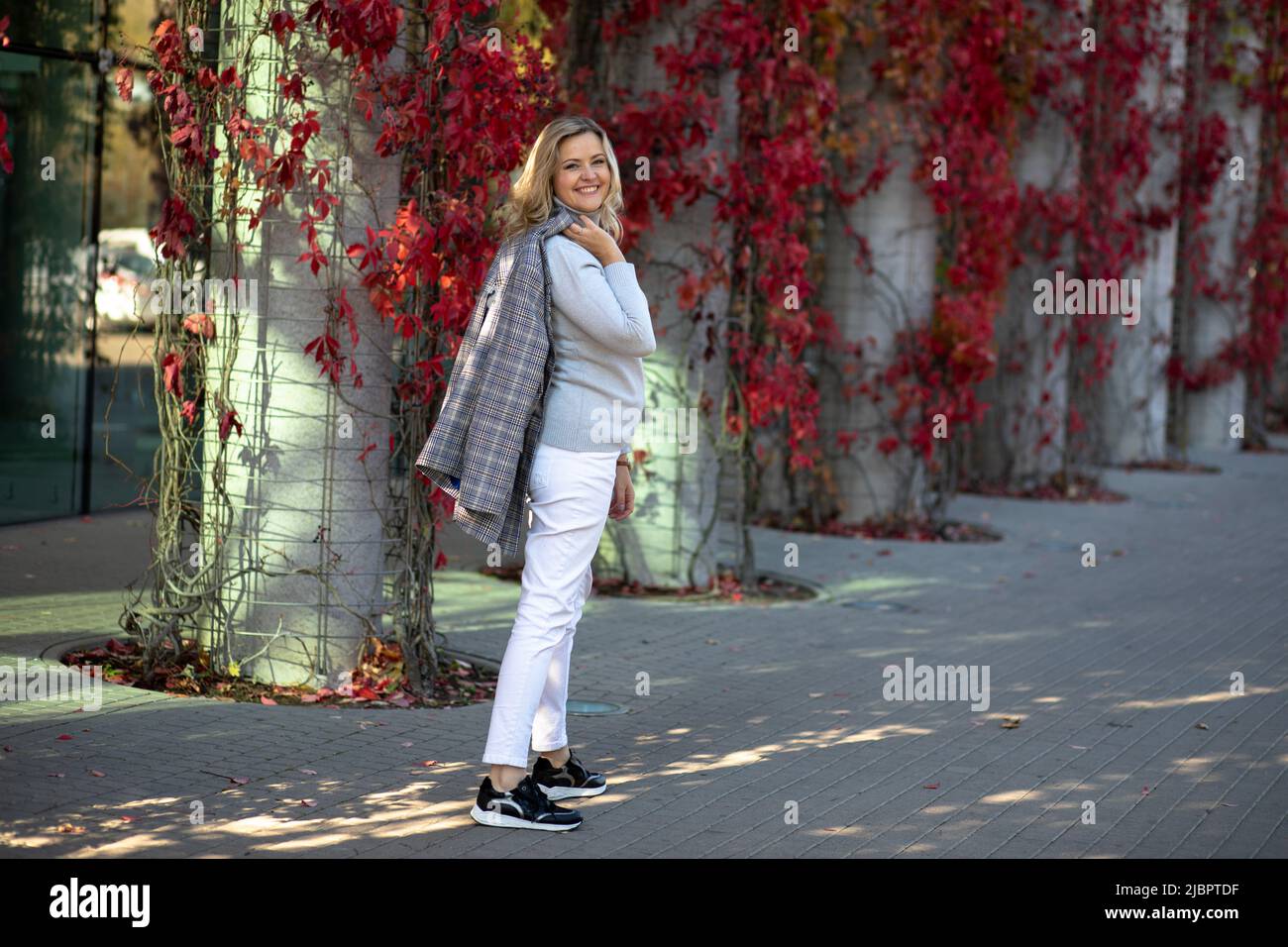 Bellamente vestido rubio rubio de 35-40 años de edad en pantalones blancos y chaqueta ligera poses, sonriendo a la cámara en la calle de la ciudad con follaje rojo brillante Foto de stock