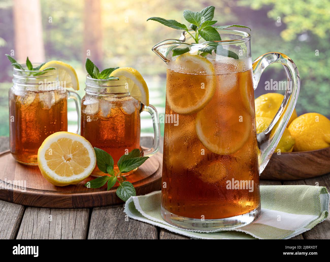 Jarra de té frío helado con menta, rodajas de limón y hielo con dos vasos o té en una mesa de madera y fondo rural de verano Foto de stock