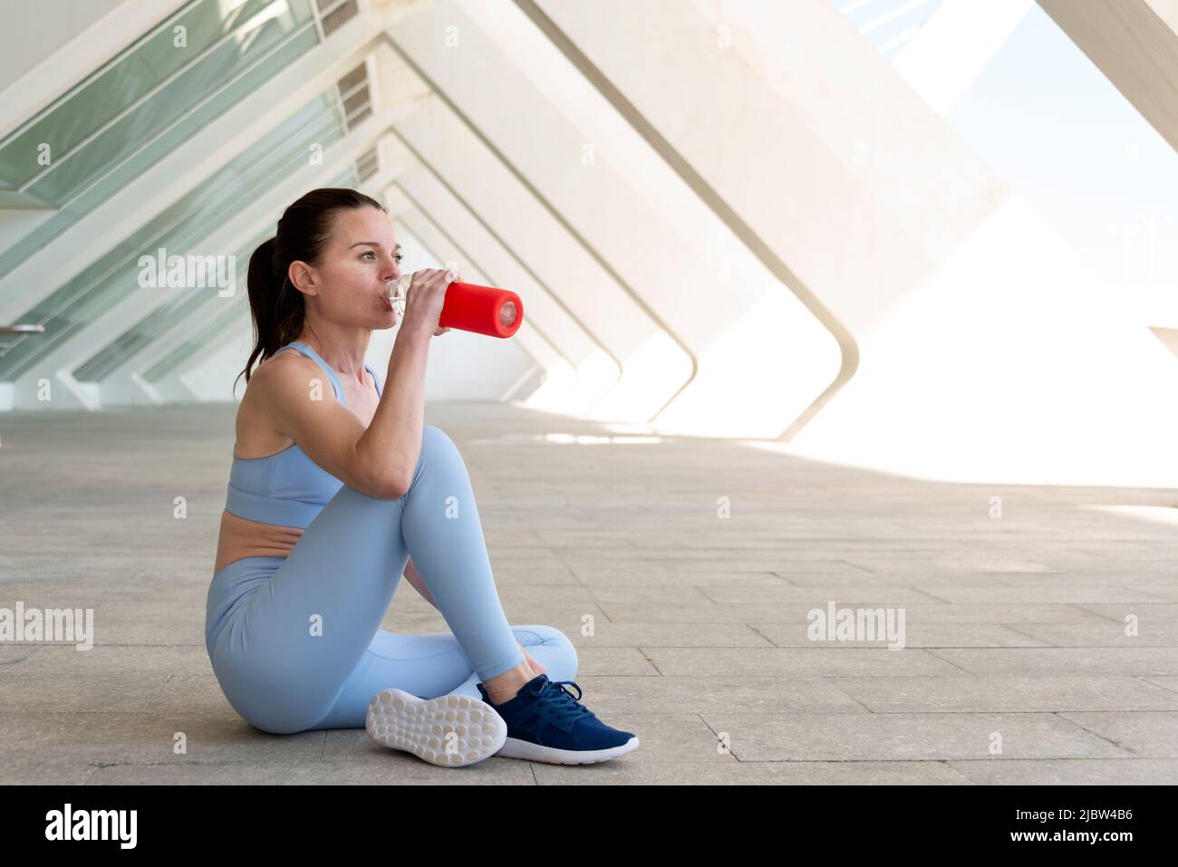 Mujer deportiva y en forma sentada bebiendo agua de una botella de vidrio después de hacer ejercicio o correr. Fondo urbano. Foto de stock
