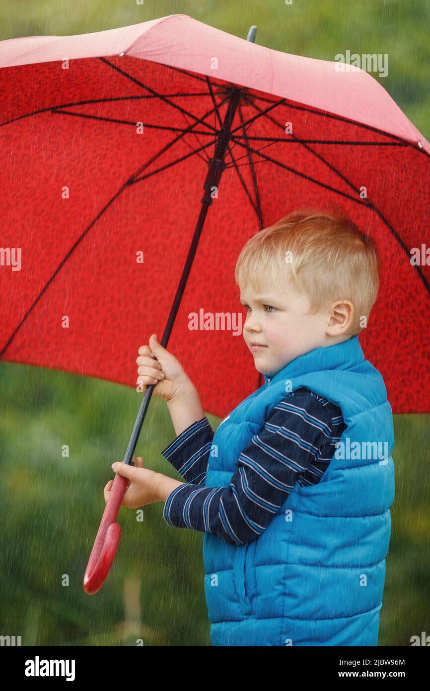 Adorable niño sosteniendo un paraguas rojo durante una tormenta de lluvia. Foto vertical. Foto de stock
