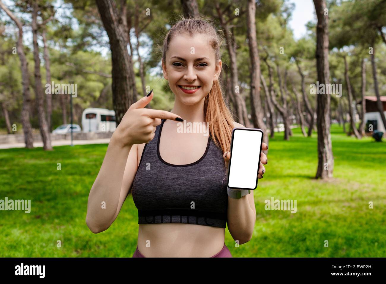 Mujer joven sonriente segura con sujetador deportivo parado en el parque de  la ciudad, al aire libre apuntando con el dedo a un teléfono móvil con  pantalla blanca en blanco. Gimnasio Fotografía