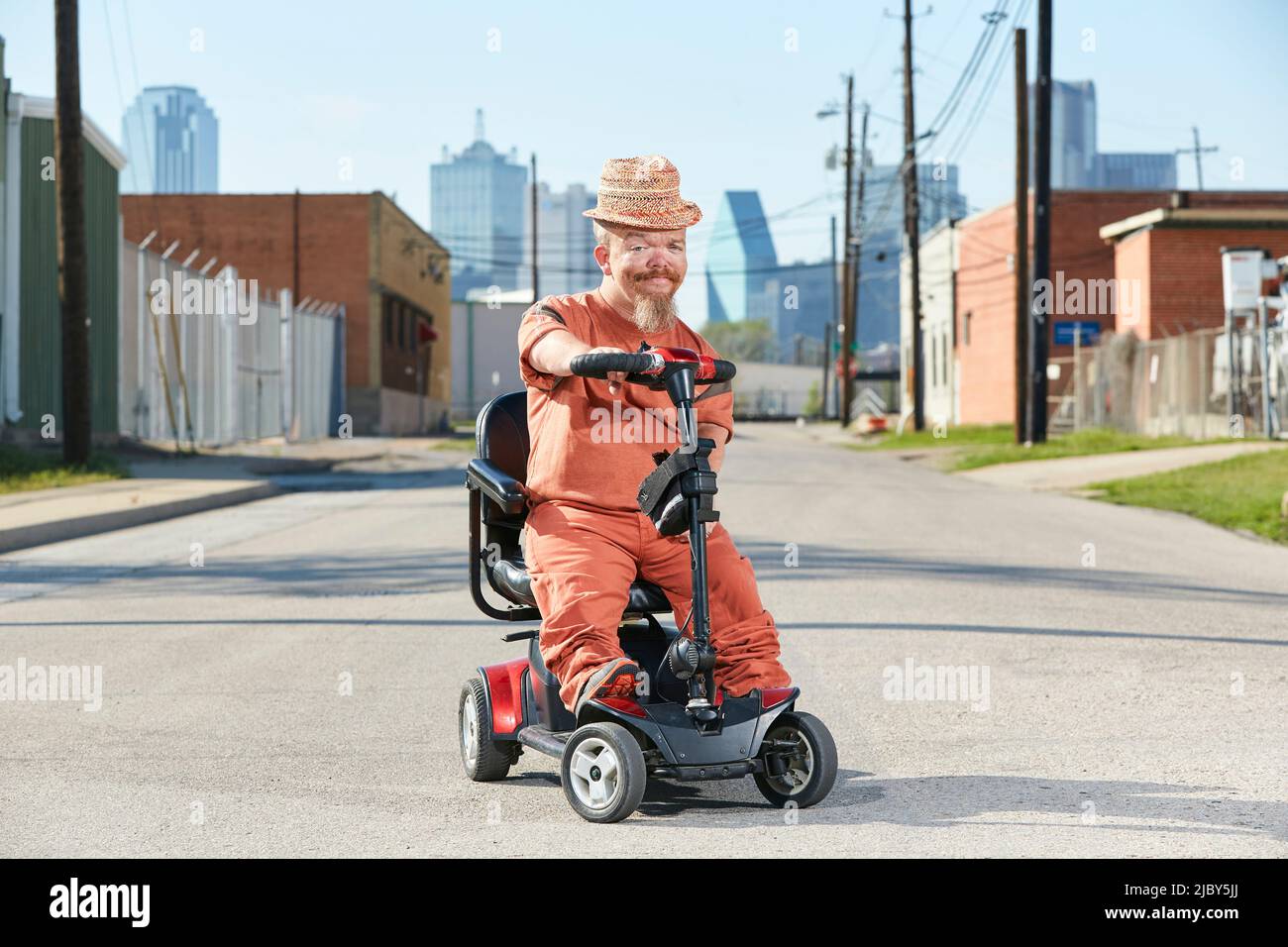 Retrato de un adulto caucásico pequeño en un scooter de movilidad aparcado en medio de la calle con el horizonte de Dallas Texas en el fondo. Foto de stock