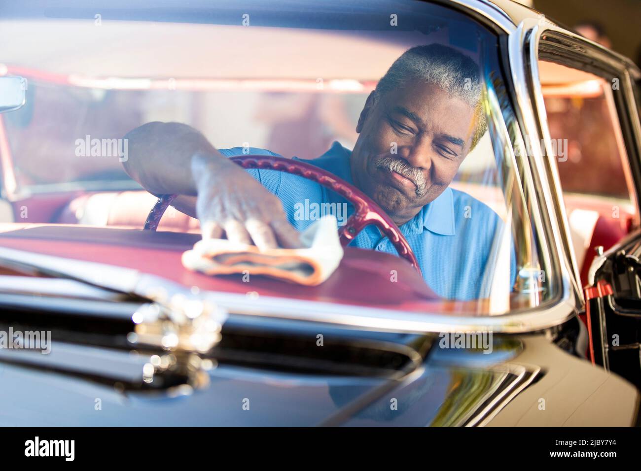 Hombre mayor sentado en el asiento del conductor de 1960 Oldsmobile Super 88 Holiday Sport Sedan, limpiando el salpicadero con un paño para el polvo Foto de stock