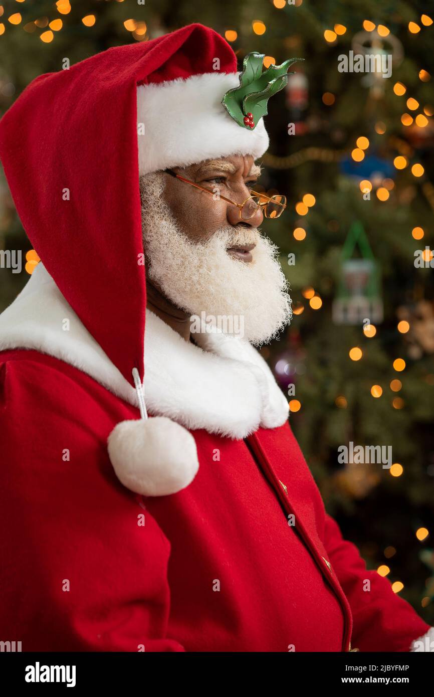 Un hombre afroamericano vestido como Papá Noel sentado frente a un árbol de Navidad se volvió perfil, mirando fuera de la cámara. Foto de stock