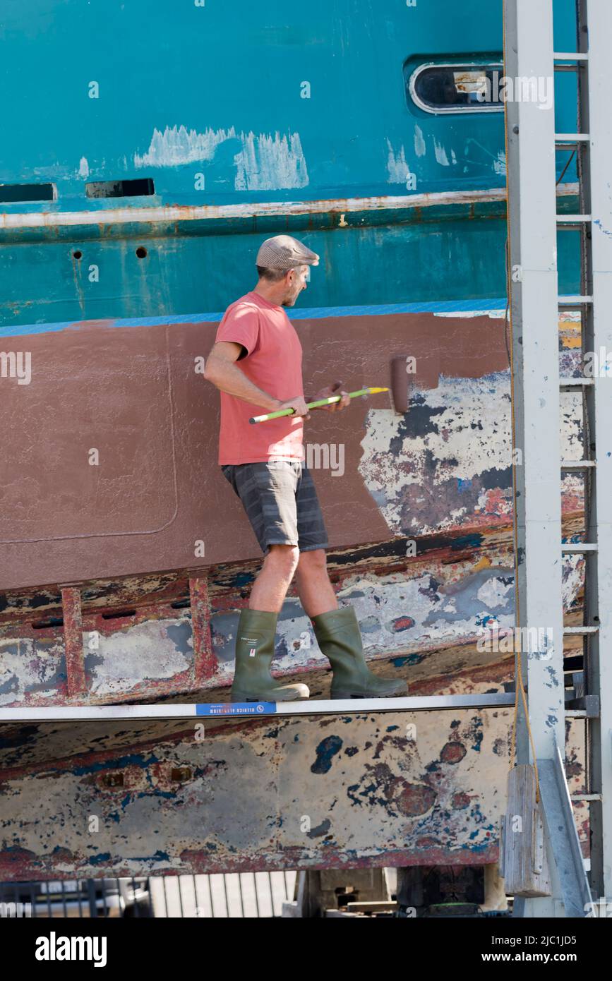 Un hombre de pie sobre una plancha de aleación utilizando un rodillo para pintar el lado de un gran arrastrero de pesca en una rampa Foto de stock