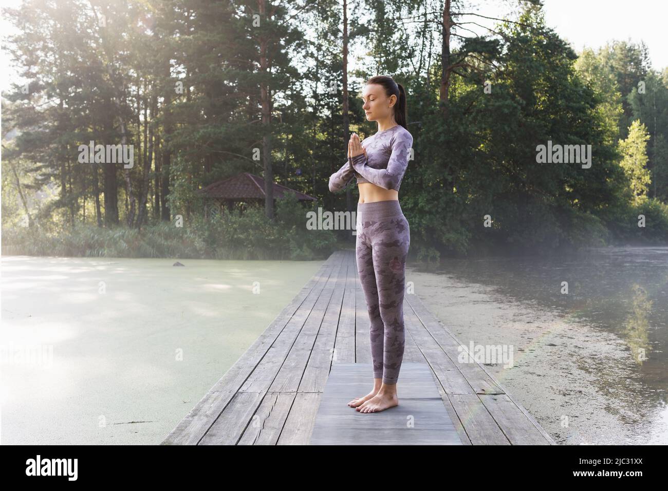 Una mujer practicando yoga, haciendo una variación del ejercicio vrikshasana, pose de árbol con namaste, de pie en un puente de madera en una mañana de verano en el Foto de stock