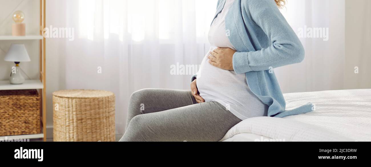 La mujer que espera a un bebé con amor y ternura acaricia su barriguita embarazada mientras está sentada en la cama. Foto de stock