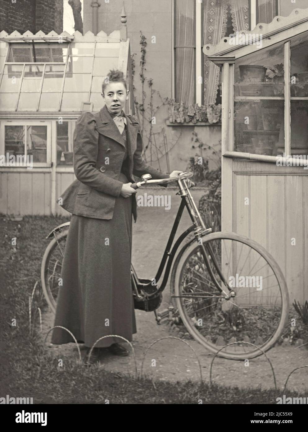 Una mujer posa con la bicicleta de su dama en su jardín trasero, Reino Unido c. 1900. Lleva ropa adecuada para montar a caballo. El jardín trasero alberga dos invernaderos. Esto es de un negativo de vidrio victoriano antiguo – una fotografía vintage de 1800s/1900s. Foto de stock