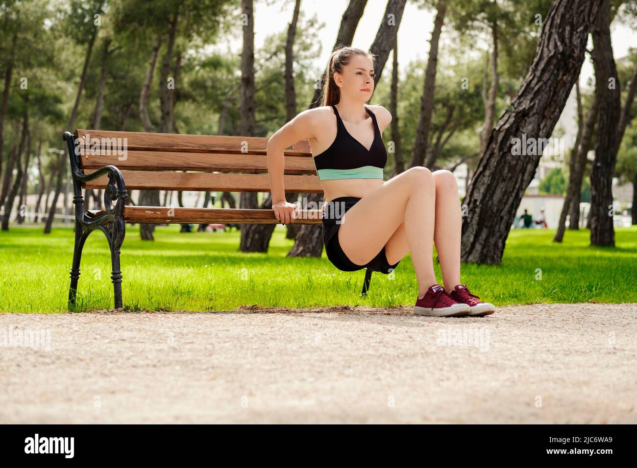 Una mujer caucásica linda que usa ropa deportiva en el parque de la ciudad,  al aire libre, concentrándose en su entrenamiento, apoyándose en un banco  del parque, haciendo ejercicios de tríceps Fotografía