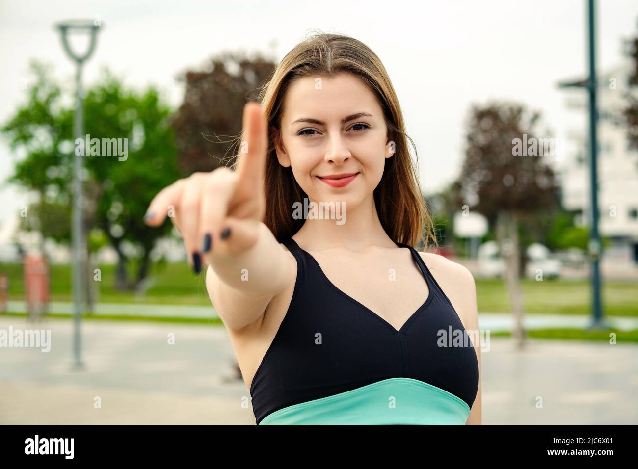Retrato de una mujer alegre con sujetador deportivo negro parado en el parque de la ciudad, al aire libre mostrando y señalando con los dedos número uno mientras sonriendo conf Foto de stock