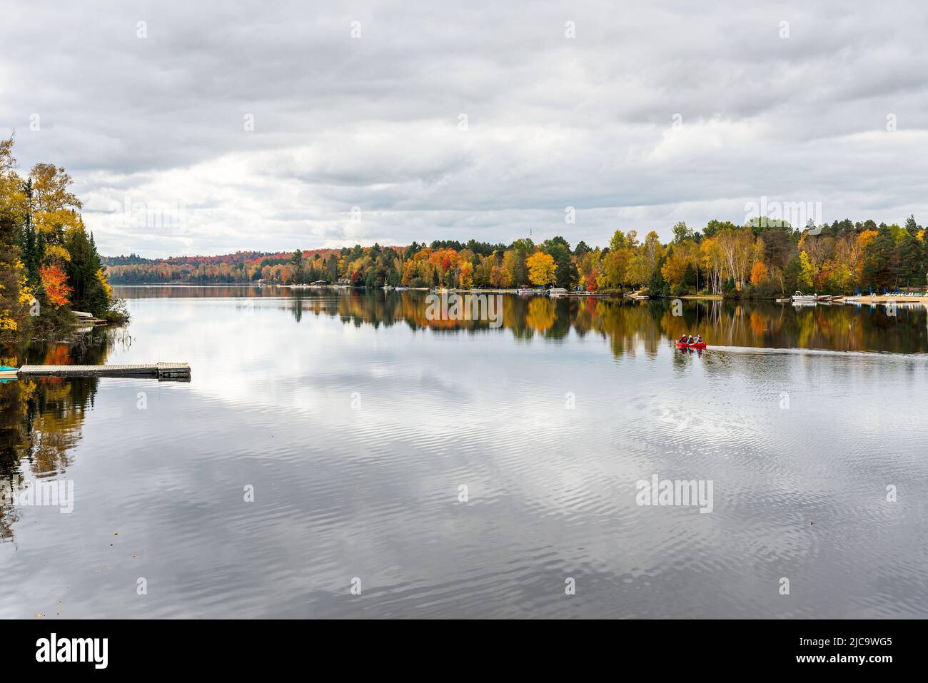 Hermoso lago con orillas boscosas en el pico del follaje otoñal en un día nublado de otoño. La gente en una canoa está en el medio del lago Foto de stock