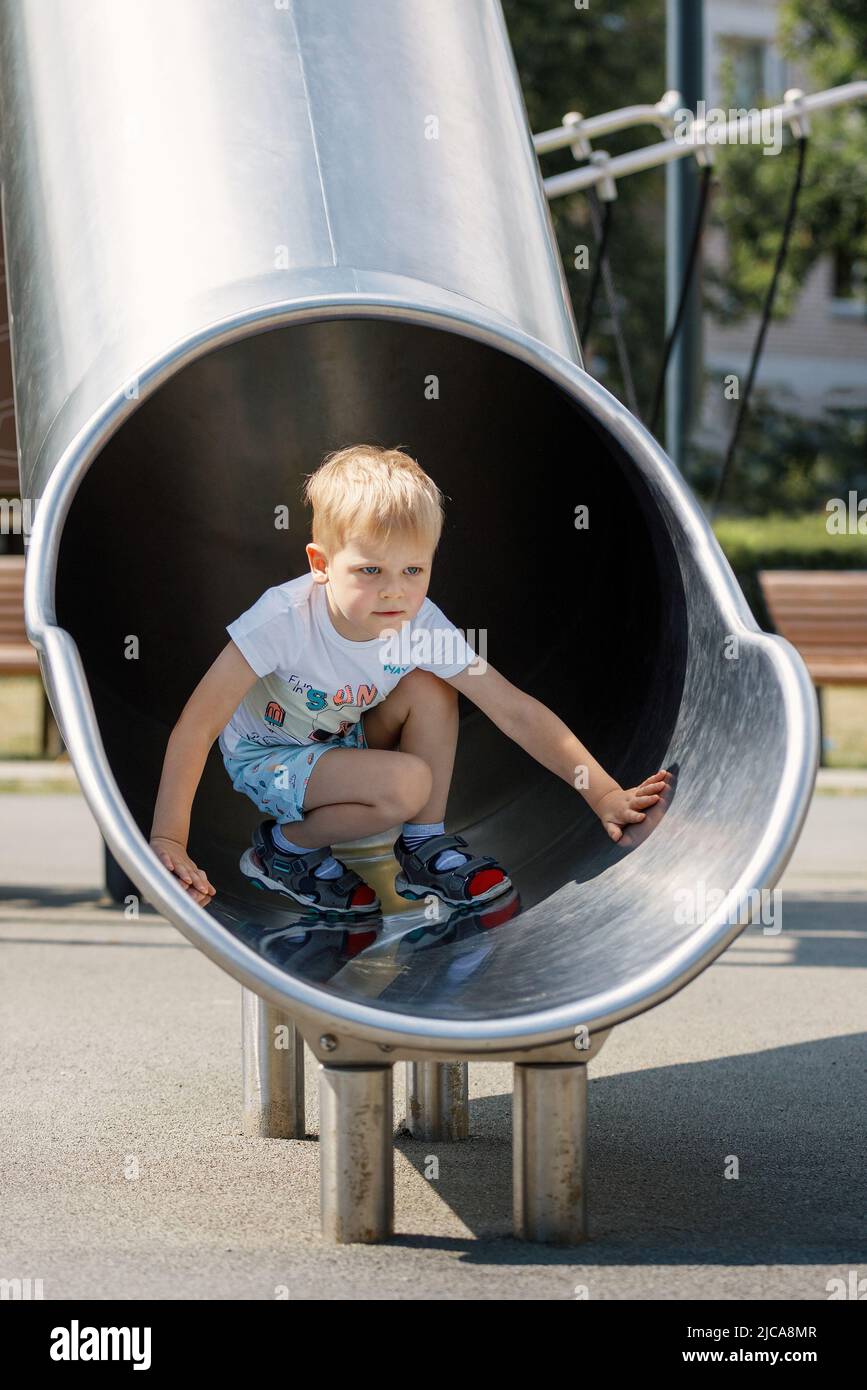 Niño alegre deslizándose en tobogán de tubo en el patio de juegos. Jardín de infantes para niños de edad elemental. Foto de stock