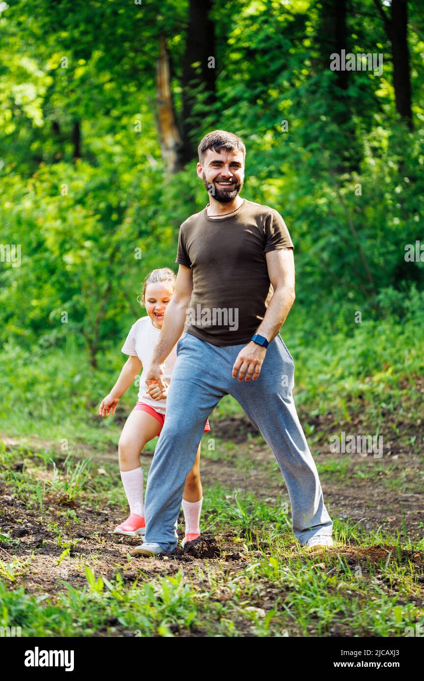 Retrato de familia feliz caminando en el bosque del parque alrededor de los árboles, divertirse. Hija pequeña manteniendo apretado el control del padre. Foto de stock