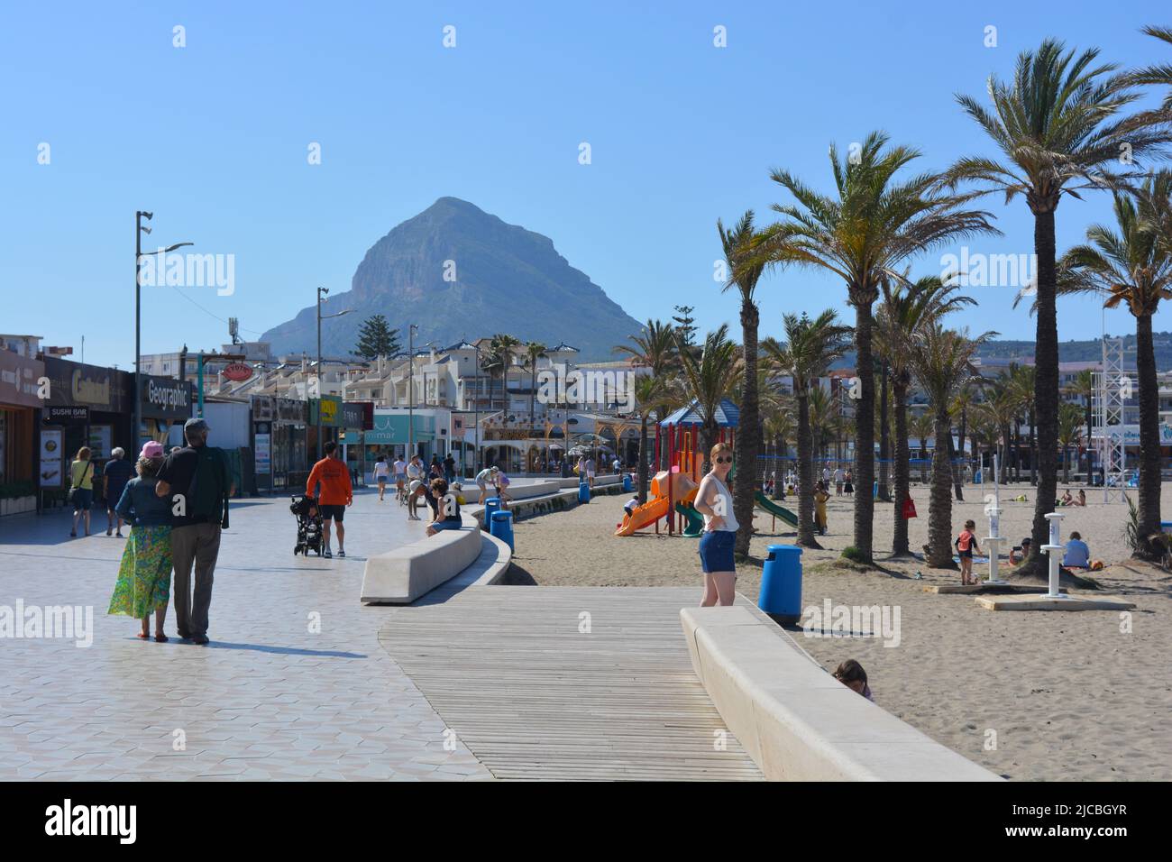 Javea, Alicante, España - Mayo 11 2022: Playa Arenal con turistas, tiendas y restaurantes en la orilla del mar y Montgo en el fondo Foto de stock