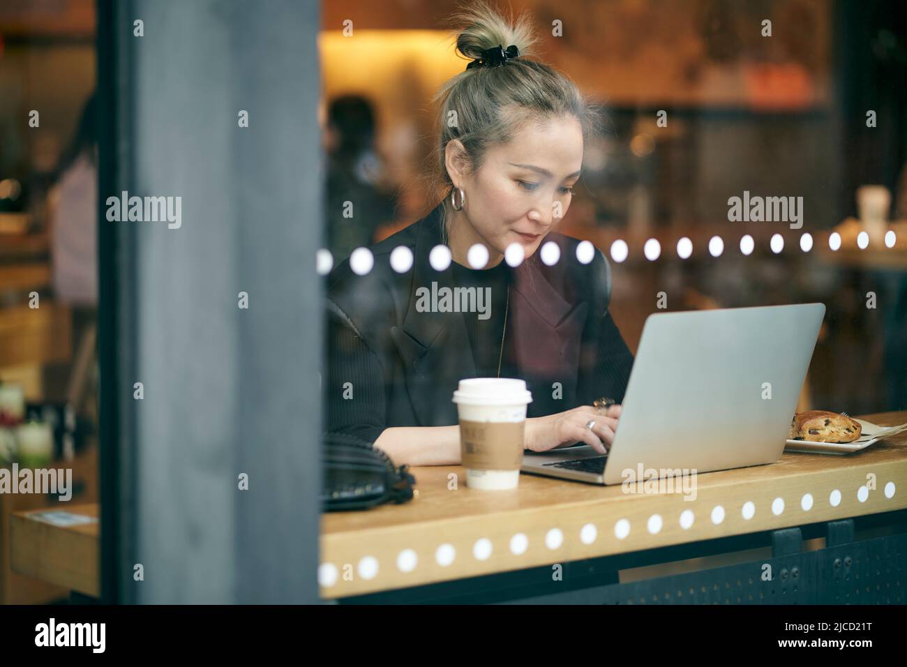 a través de una toma de cristal de una mujer de negocios asiática de mediana edad que trabaja en la cafetería utilizando un ordenador portátil Foto de stock