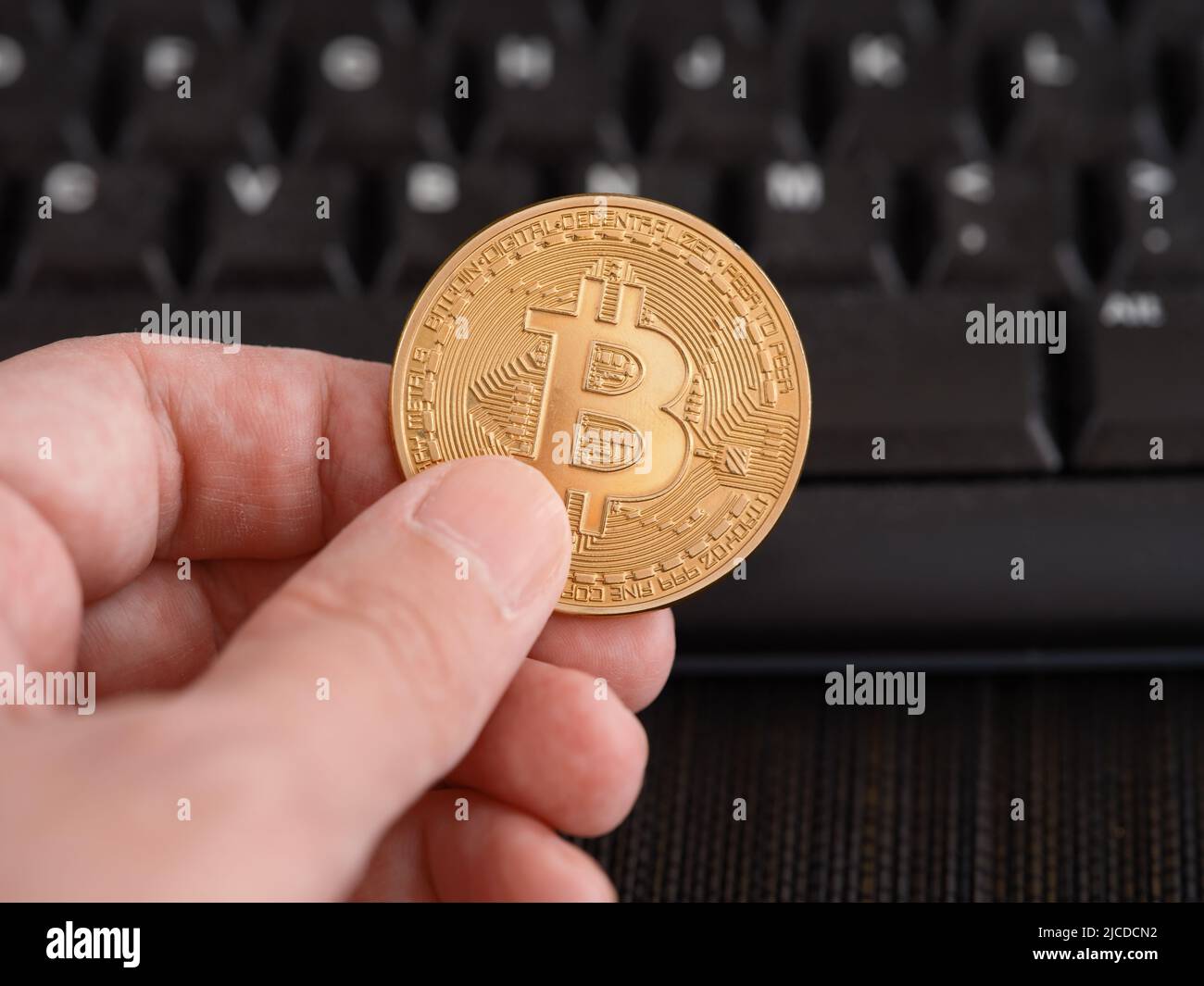 Un hombre sosteniendo una moneda de bitcoin dorada en su mano contra un teclado de ordenador. Primer plano. Foto de stock