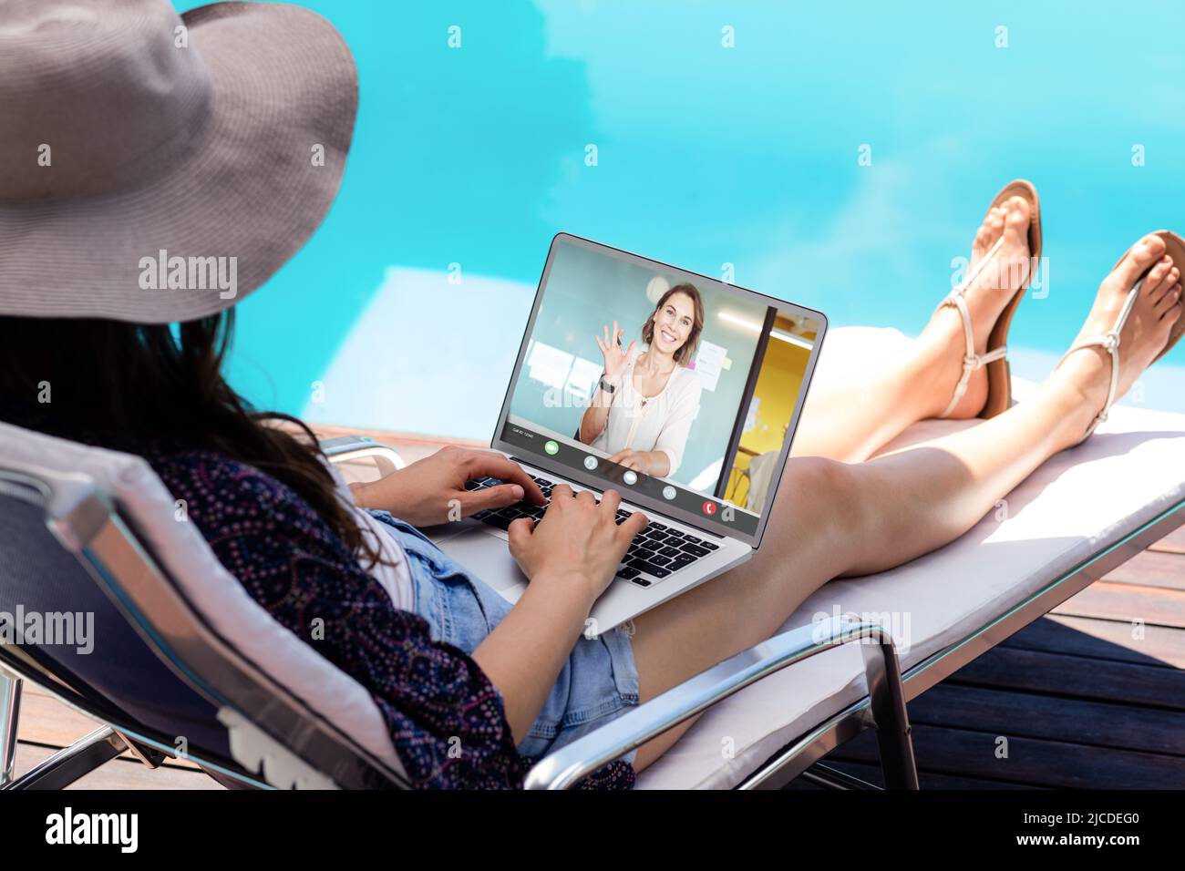Mujer de negocios caucásica hablando con un compañero de trabajo con el ordenador portátil mientras se sienta junto a la piscina de vacaciones Foto de stock