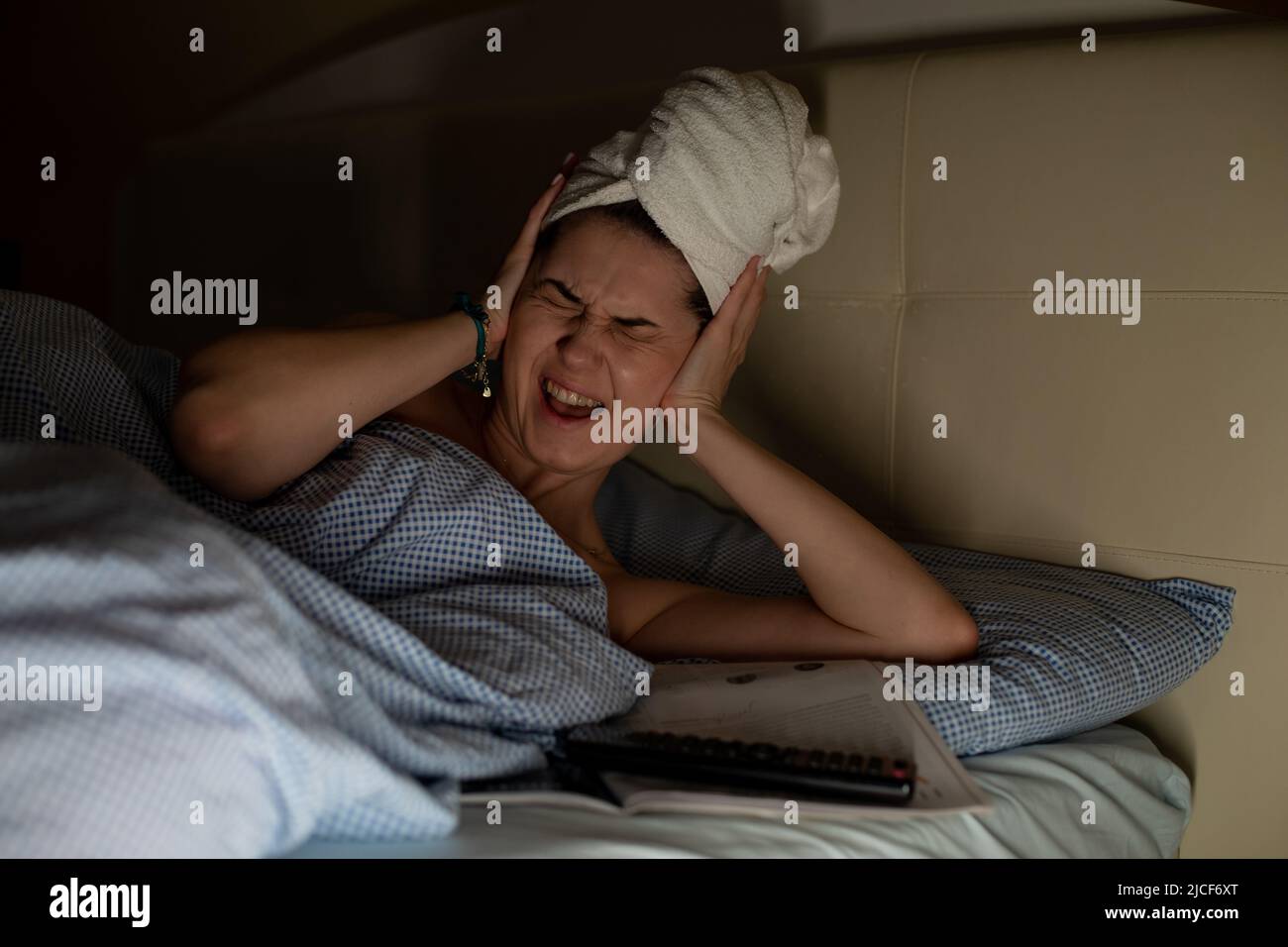 Mujer joven con toalla blanca envuelta alrededor de la cabeza tumbada en cama que sufre de dolor de cabeza, dolor de oído que cubre los oídos con las manos. Foto de stock