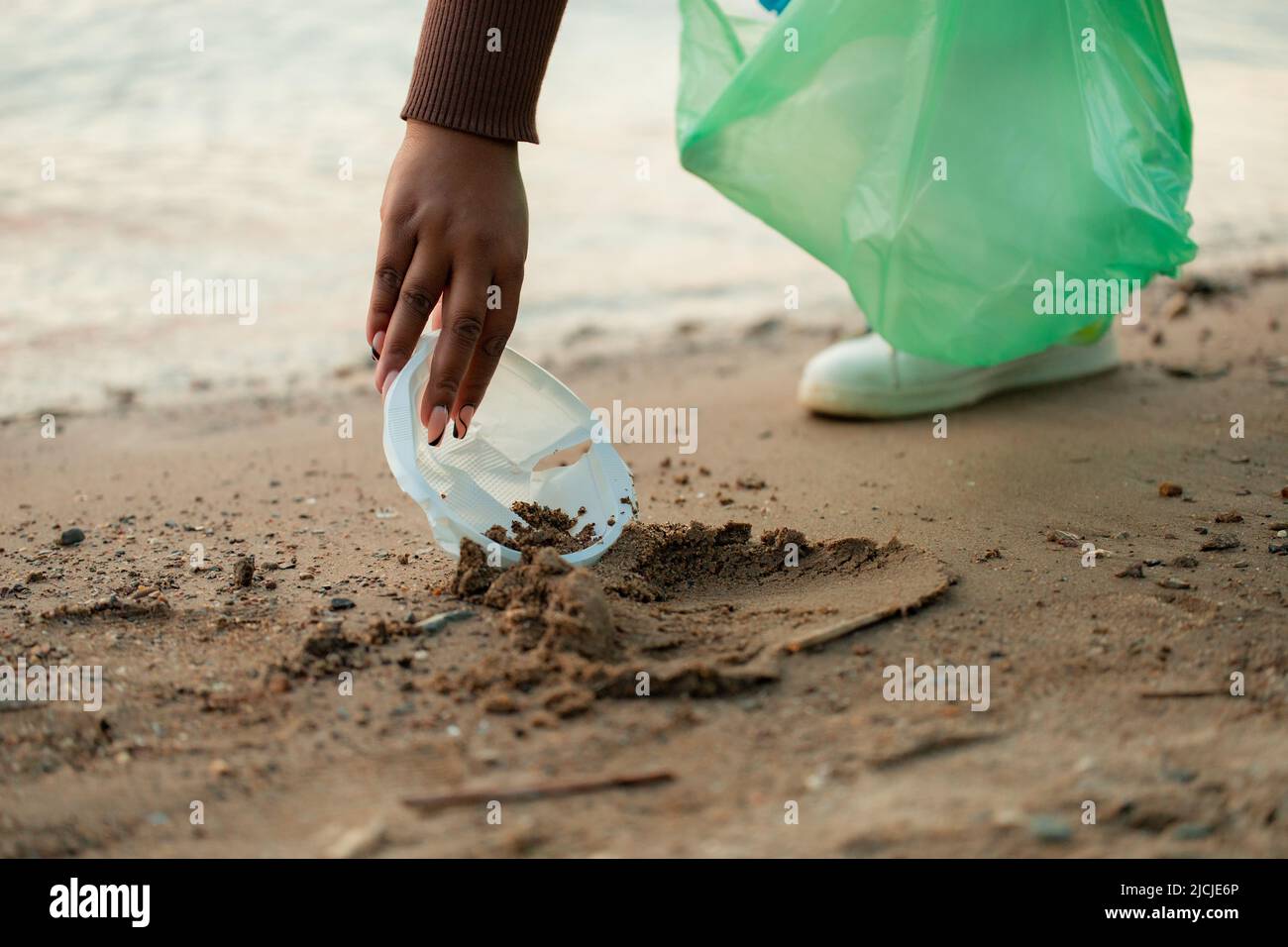 Foto recortada de una mujer africana recolectando basura derramada de la arena en la playa en bolsa de plástico verde. Ecología, contaminación. Foto de stock