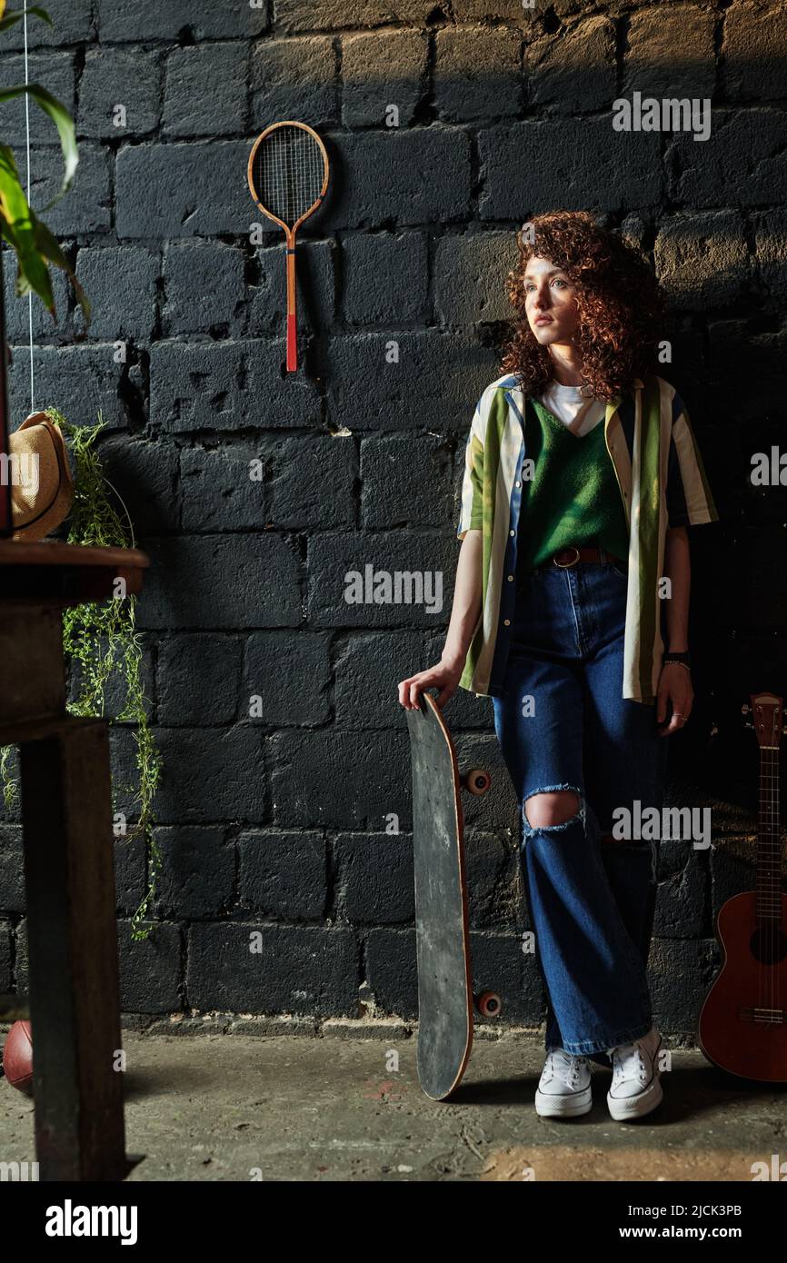 Chica serena con skateboard de pie delante de la cámara contra la pared de  ladrillo gris con raqueta de tenis en el garaje o estudio moderno  Fotografía de stock - Alamy