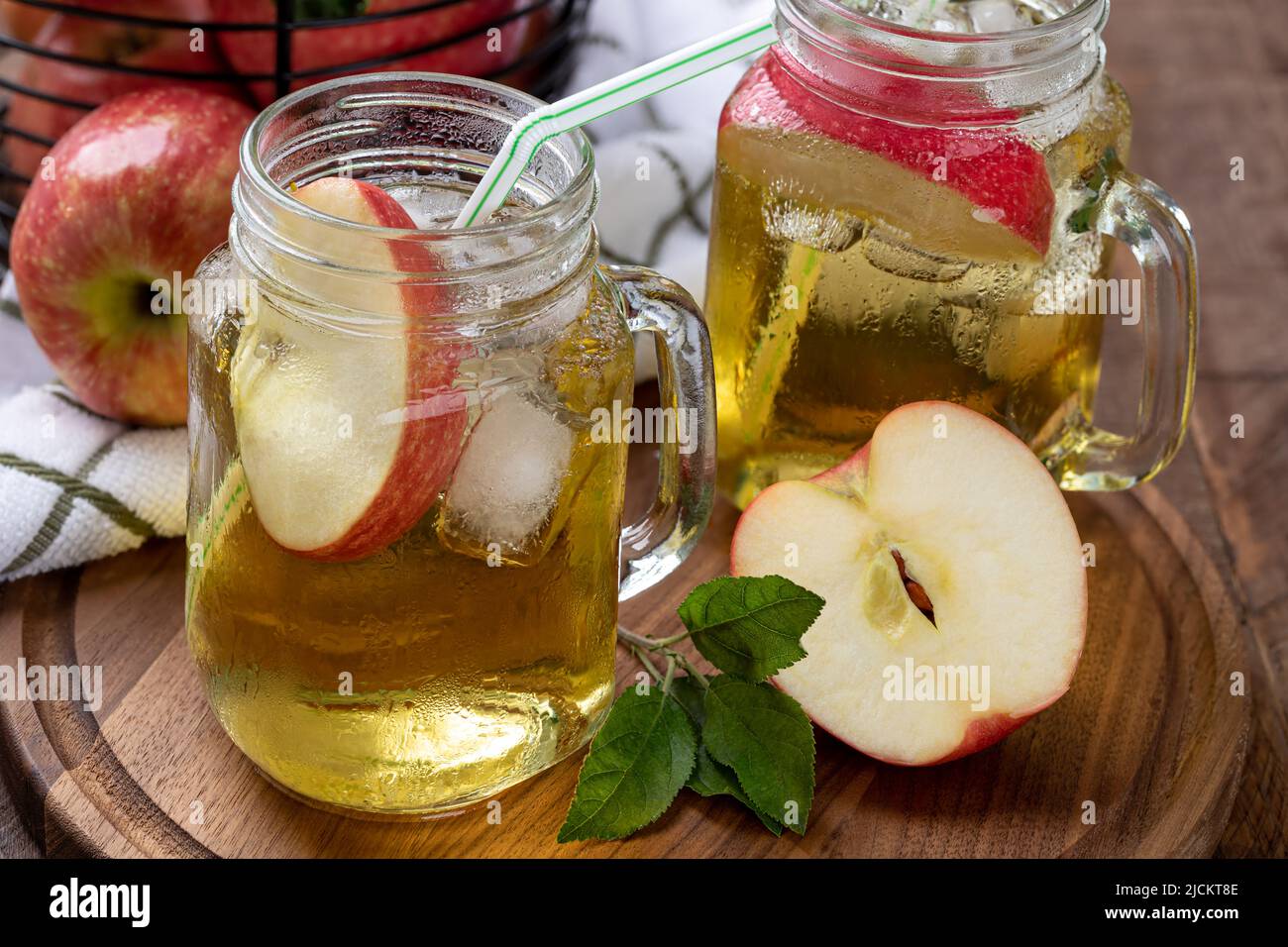 Dos vasos de zumo de manzana con rodajas de manzana y hielo en un plato de madera con una cesta de manzanas de fondo Foto de stock