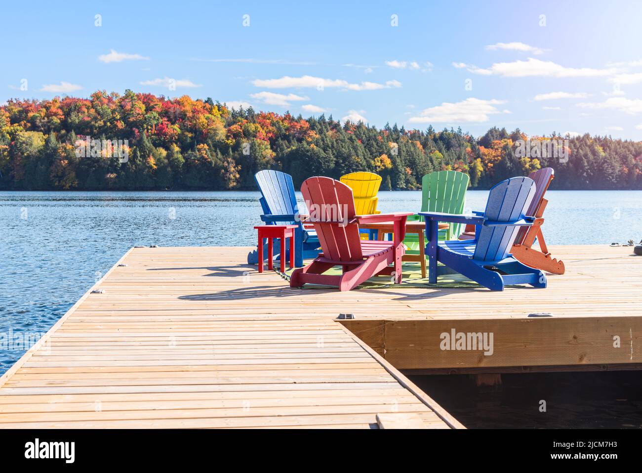 Coloristas sillas Adirondack alrededor de una mesa en un embarcadero de madera en un lago en un claro día de otoño. Foto de stock
