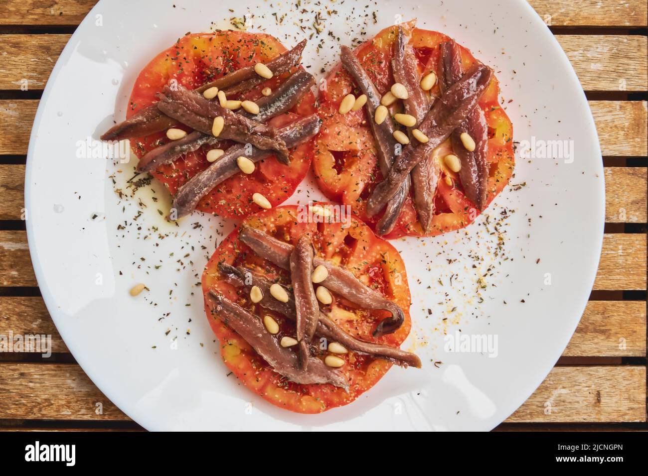 Tres rodajas de tomates cultivados orgánicamente con anchoas, piñones y especias sobre un plato blanco sobre una mesa de teca Foto de stock