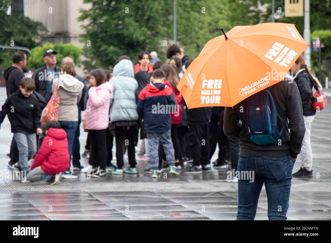 Belgrado, Serbia - 28 de mayo de 2022: Guía de viaje de pie bajo paraguas naranja con señal de excursión a pie gratuita, en una plaza de la ciudad en un día lluvioso, vista posterior y. Foto de stock
