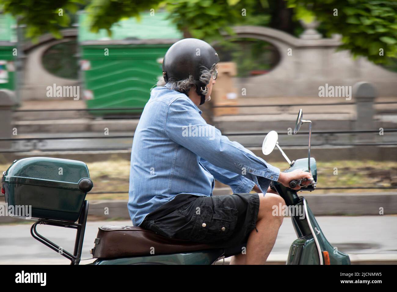 Belgrado, Serbia - 28 de mayo de 2022: Hombre de mediana edad que monta en una vespa verde scooter con la caja trasera del maletero en la calle de la ciudad junto al parque Foto de stock