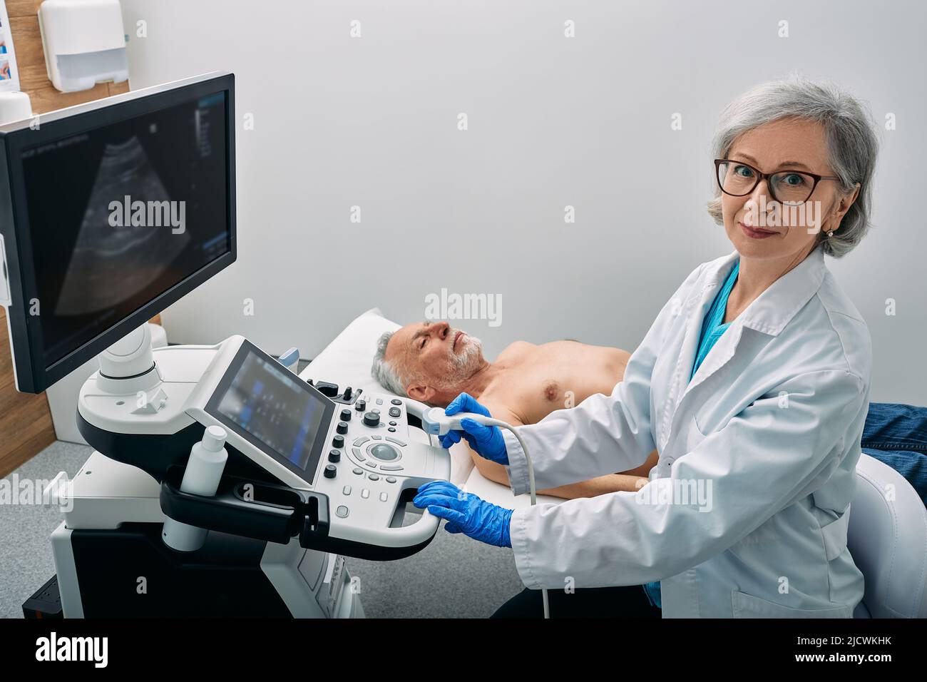 Ocupación ecografista. Retrato del ecografista cerca de la máquina de ultrasonido en la clínica médica durante la ecografía corporal del paciente masculino Foto de stock