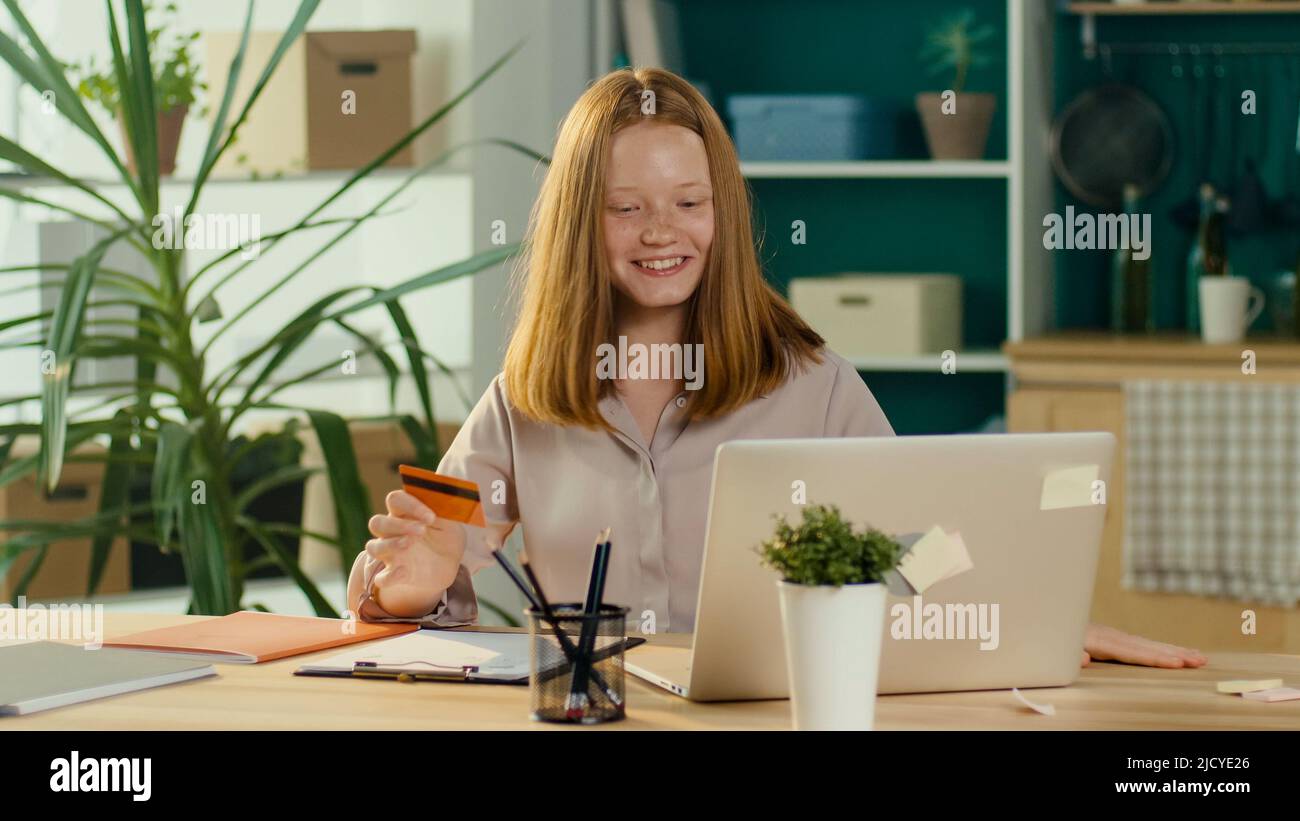 Feliz chica adolescente de pelo rojo de compras en línea con una tarjeta de la casa. Consumidor. Foto de stock