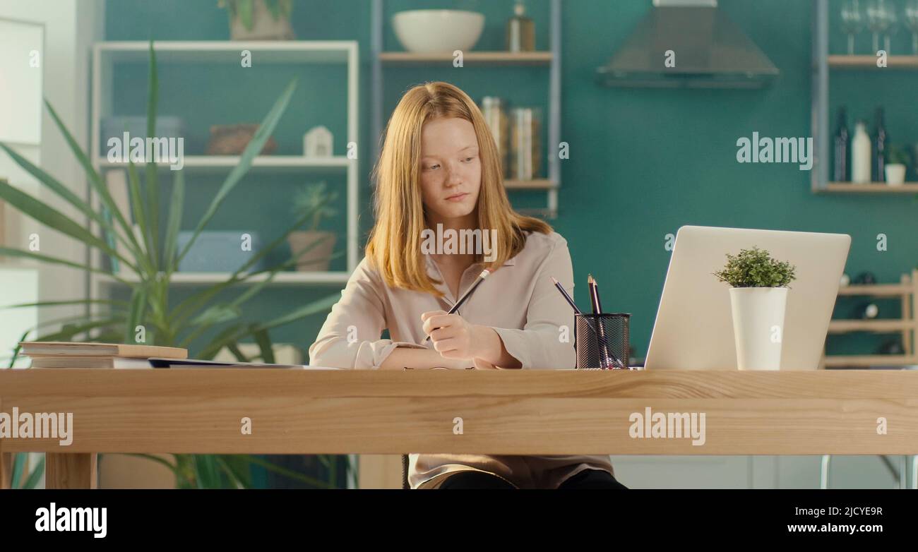 Niña adolescente de pelo rojo estudiando en casa en línea, viendo un tutorial en vídeo. Foto de stock