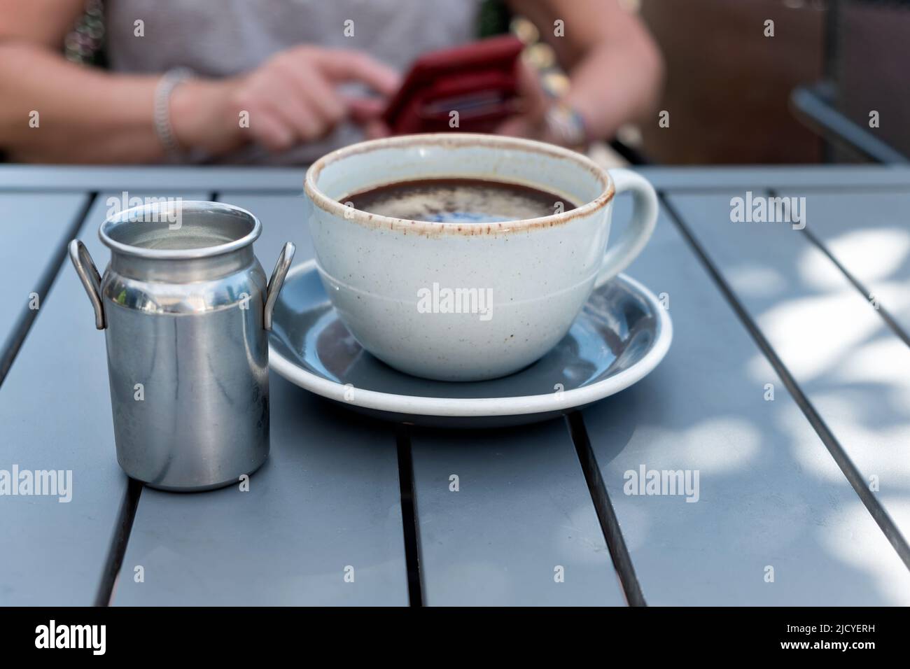 Una mujer está ocupada enviando mensajes de texto en su móvil mientras espera una taza de café recién hecha para enfriar. Ella está sentada en una mesa de café al aire libre en una noche de verano Foto de stock