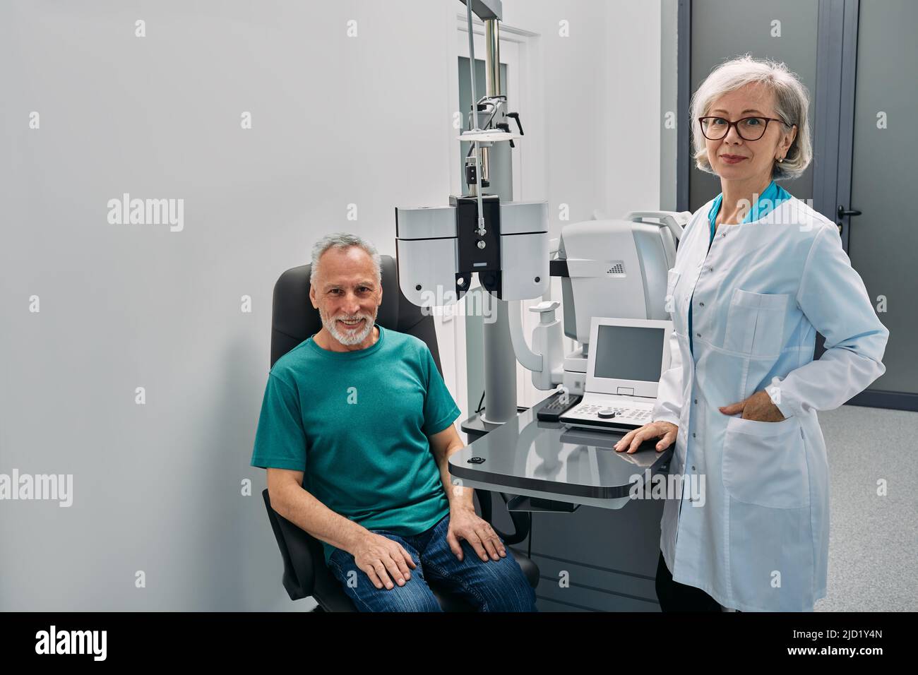 Optometrista positivo con su paciente masculino mayor mirando amistoso a la cámara mientras consulta oftalmológica cerca de equipo oftálmico Foto de stock