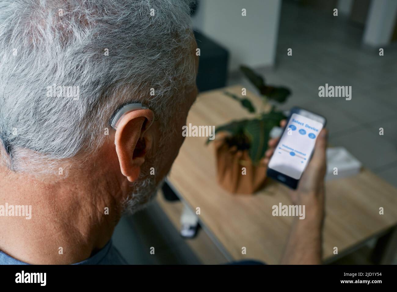 Un hombre mayor con problemas auditivos que ajusta la configuración de su audífono retroauricular mediante un smartphone. Audífonos con tecnologías innovadoras en audiología Foto de stock