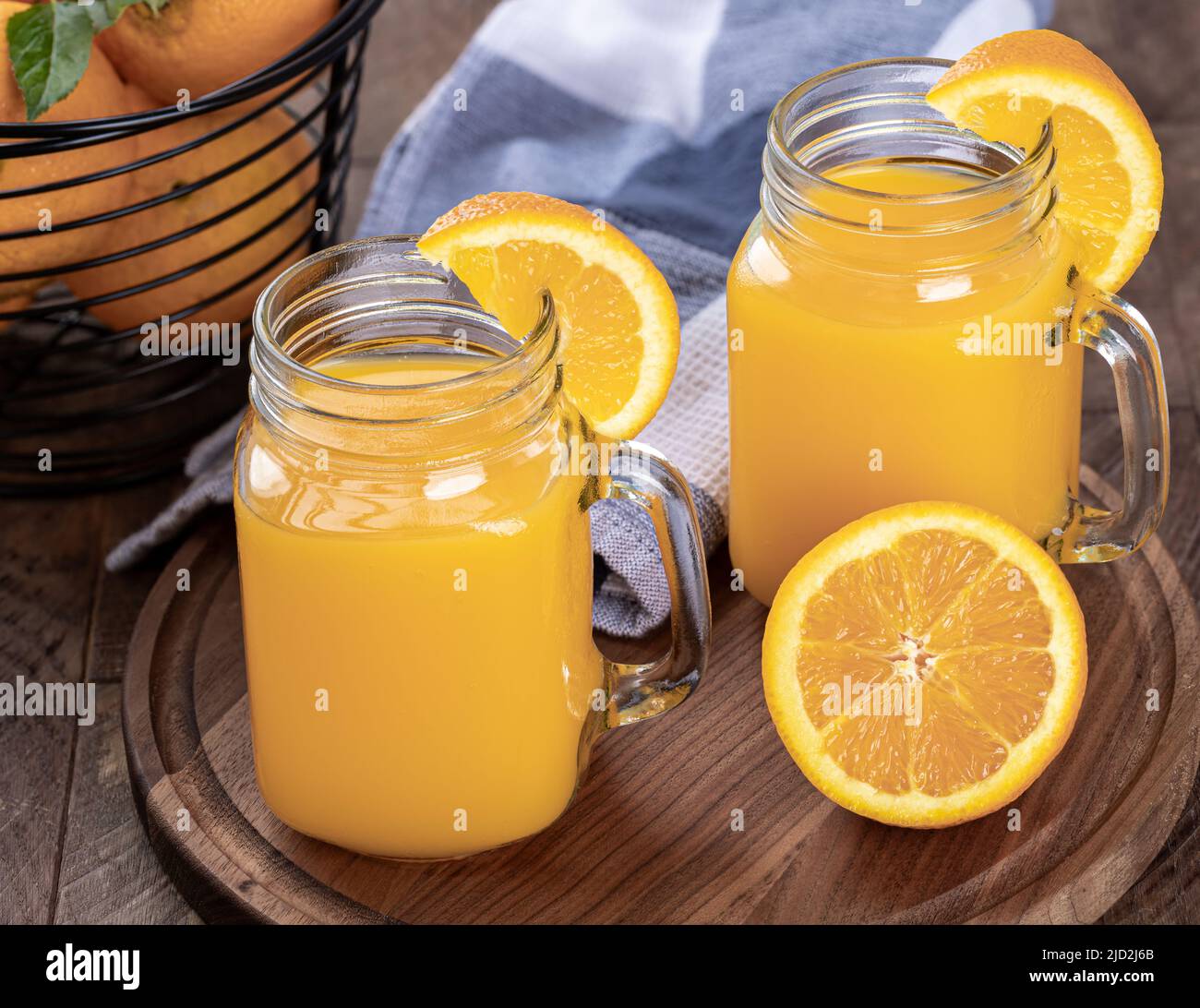 Dos vasos de zumo de naranja con siceses de naranja en un plato de madera con una cesta de naranjas de fondo Foto de stock