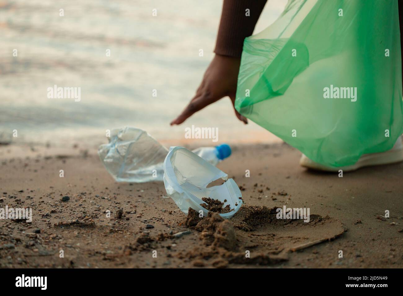 Mujer africana irreconocible que recoge basura derramada de la arena en la playa en bolsa de plástico verde. Ecología, contaminación. Foto de stock