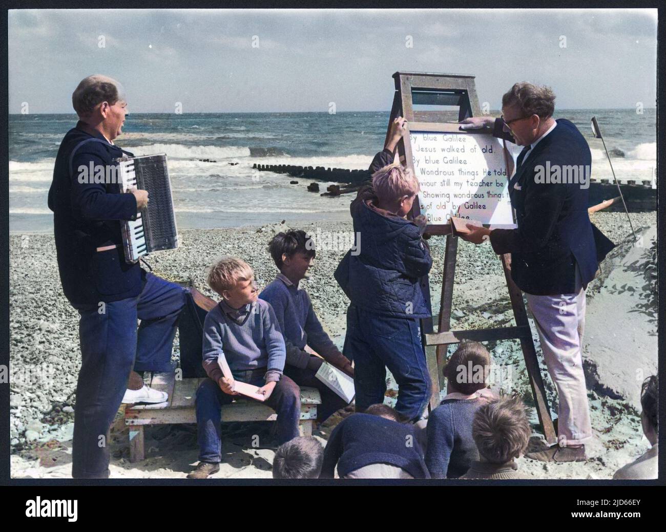 Un servicio religioso al aire libre que se realiza en una playa, con acompañamiento de acordeón para el canto, y las palabras que se muestran en una caballete. Versión coloreada de : 10007066 Fecha: Alrededor de 1965 Foto de stock