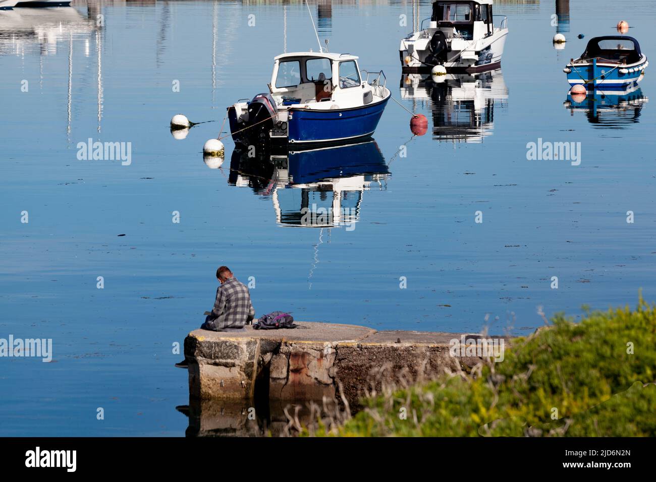 Reflexión tranquila. Un hombre sentado en un puerto con un ambiente reflexivo, con barcos que proyectan reflejos en un mar como espejo en una mañana soleada y luminosa Foto de stock