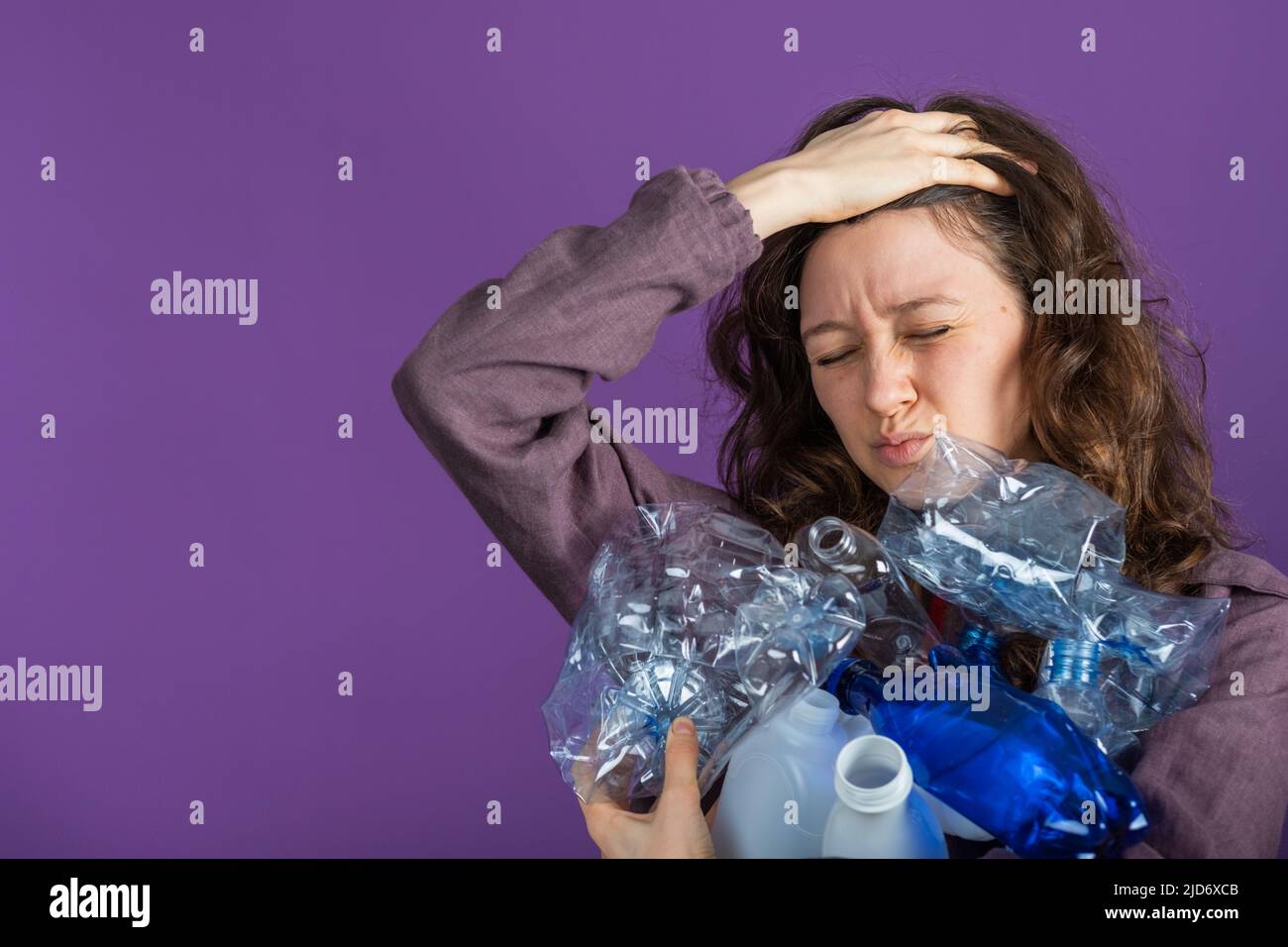 Retrato de una mujer sosteniendo un manojo de botellas de plástico en su mano y sosteniendo su cabeza con la otra mano sobre un fondo púrpura, clasificando y reciclando plástico, cuidando el medio ambiente Foto de stock