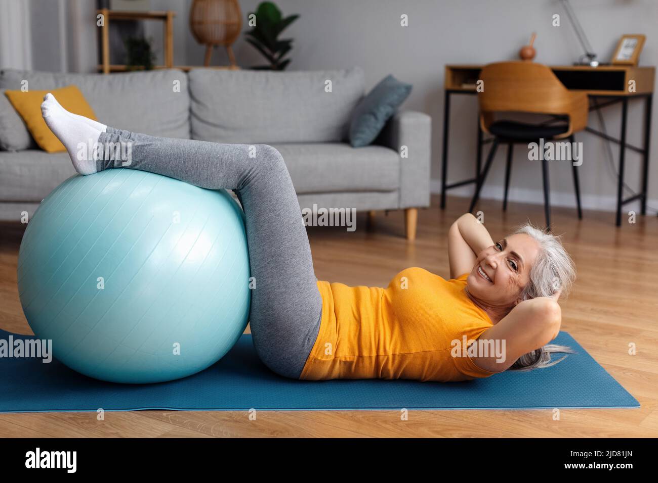 Señora mayor deportiva ejercitando sus músculos de abs con fitball en la colchoneta de yoga en casa, mirando y sonriendo la cámara Foto de stock