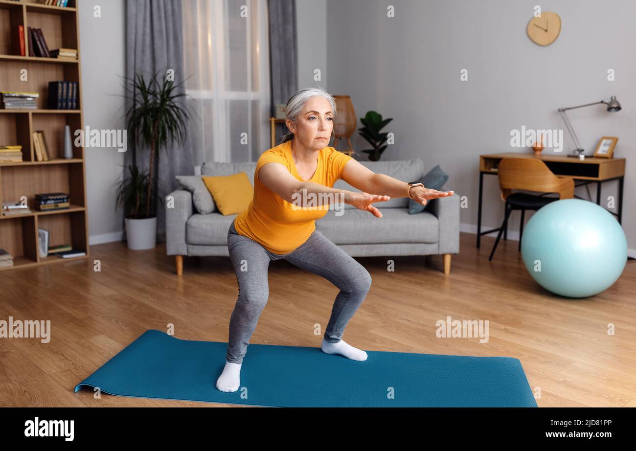 Entrenamiento en casa. Señora mayor haciendo sentadas en la colchoneta de yoga, haciendo ejercicio en casa en el interior de la sala de estar, espacio libre Foto de stock