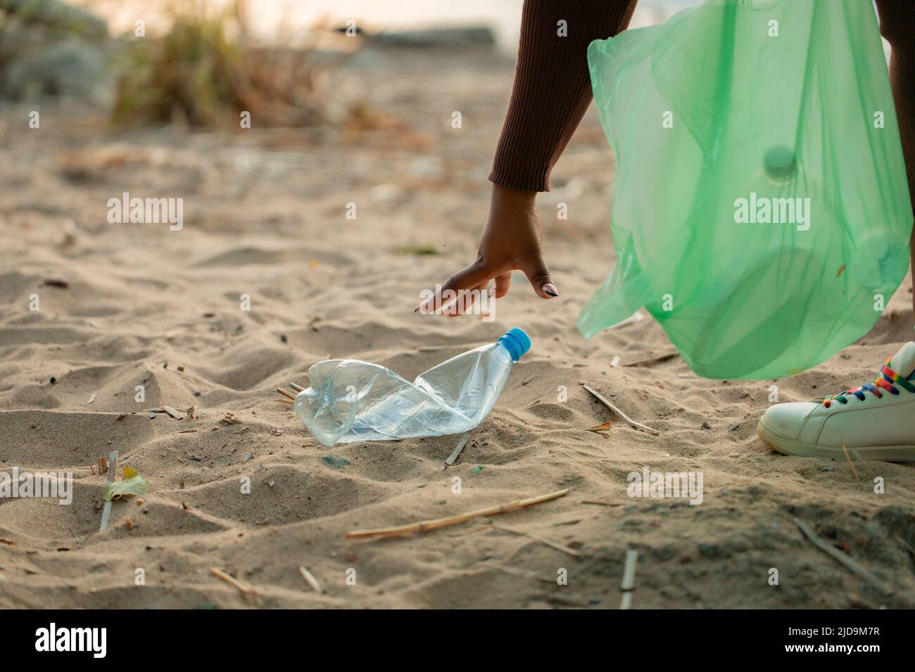 Foto recortada de una mujer africana recolectando basura derramada de la arena en la playa en bolsa de plástico verde. Ecología, contaminación. Foto de stock