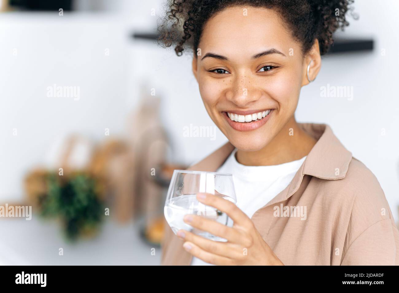 Foto de primer plano de una niña afroamericana de pelo rizado sana y positiva sosteniendo un vaso de agua limpia, cuidar la salud, controlar la tasa diaria de agua corporal, mira la cámara con una sonrisa enlozoadora feliz Foto de stock
