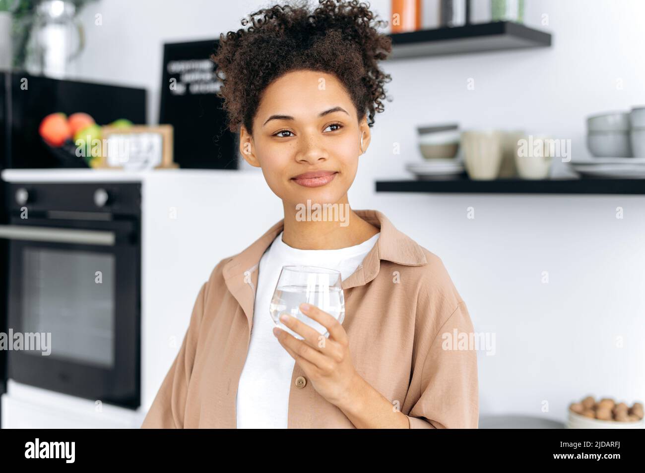 Cuidado de la salud. Foto de una sana chica afroamericana de pelo rizado que sostiene un vaso de agua limpia, cuida de la salud, controla la tarifa diaria del agua corporal, mira, sueña, sonríe Foto de stock